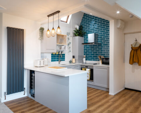 An open plan small attic flat kitchen is illuminated by thee lamps over the work area and a roof window above the sink. The cooking area is blue tiled from bench top to ceiling. A large dark grey radiator is in a vertical position to the left of the work surface. A coat is hanging on the back of the door.