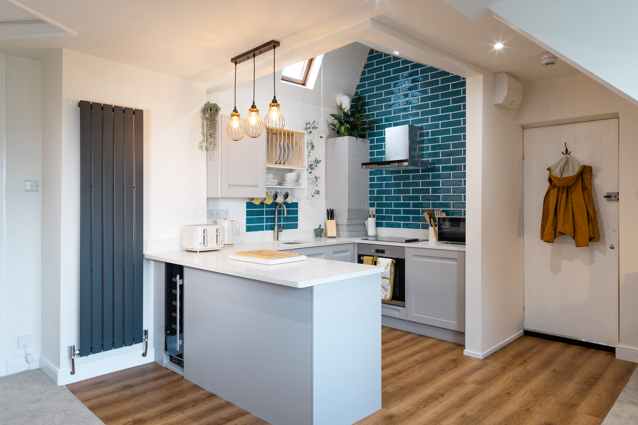 An open plan small attic flat kitchen is illuminated by thee lamps over the work area and a roof window above the sink. The cooking area is blue tiled from bench top to ceiling. A large dark grey radiator is in a vertical position to the left of the work surface. A coat is hanging on the back of the door.